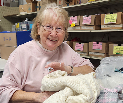An older woman with glasses and a pink sweater smiles while sorting folded clothes in what appears to be a donation center, with labeled boxes on shelves behind her.