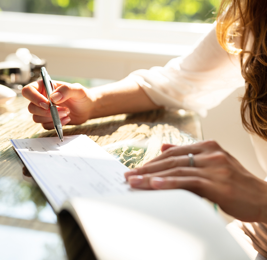 A person with long hair writes on a check with a pen while sitting at a sunlit table. Only their hands and part of their arm are visible.