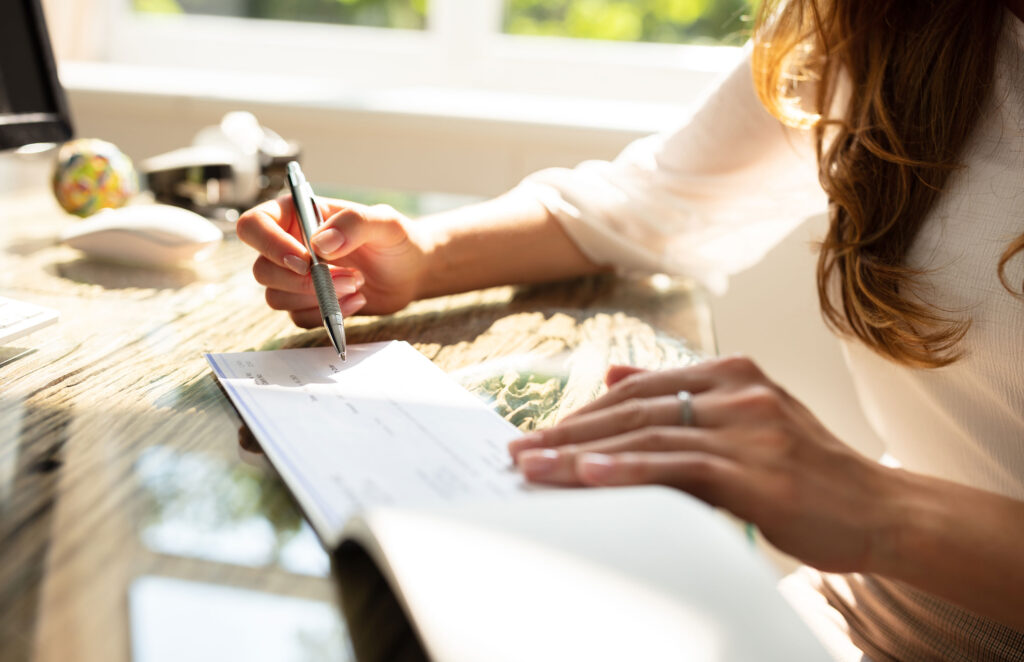 A woman sits at a sunlit desk, writing a check with a pen. Her hand holds the checkbook open, and she wears a ring on her finger. A computer and office items are blurred in the background.