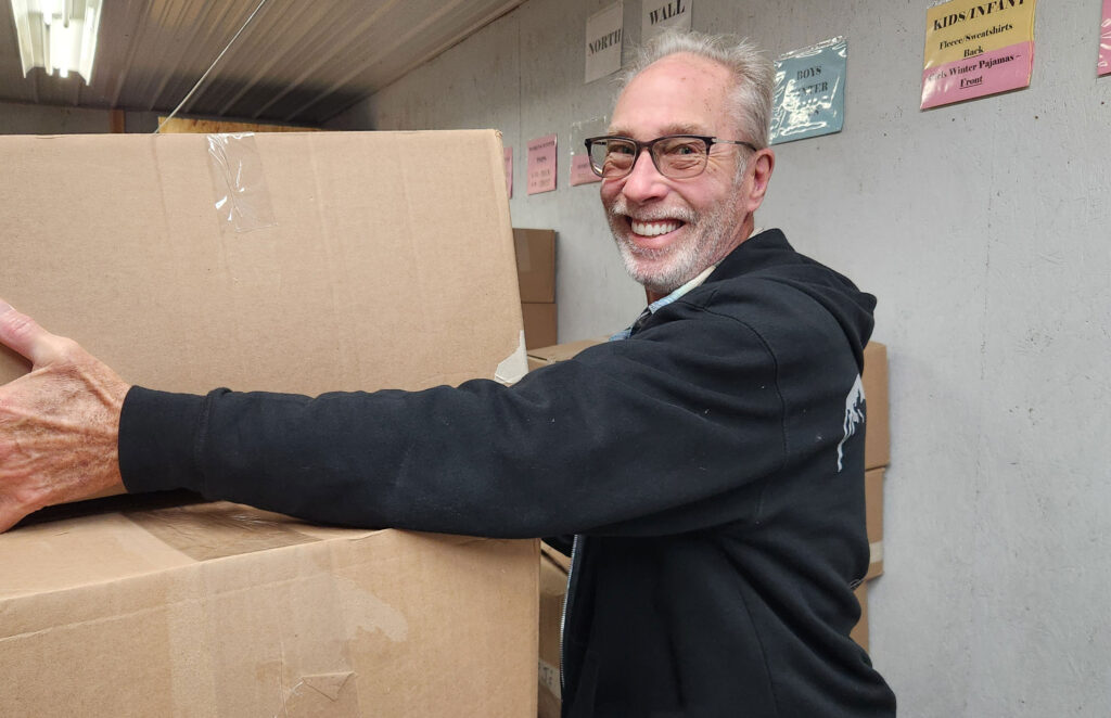 A smiling man wearing glasses and a black hoodie holds a large cardboard box in a storage room with colorful signs on the wall behind him.