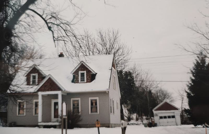 A small, gray house with white trim sits in a snowy yard, bare trees around it. A detached garage is nearby, also covered in snow, under an overcast sky.