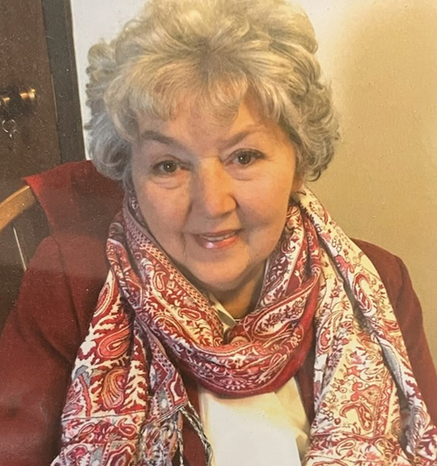 An older woman with gray curly hair smiles at the camera, wearing a red jacket and a red-and-white paisley scarf. She is seated indoors against a light-colored wall.