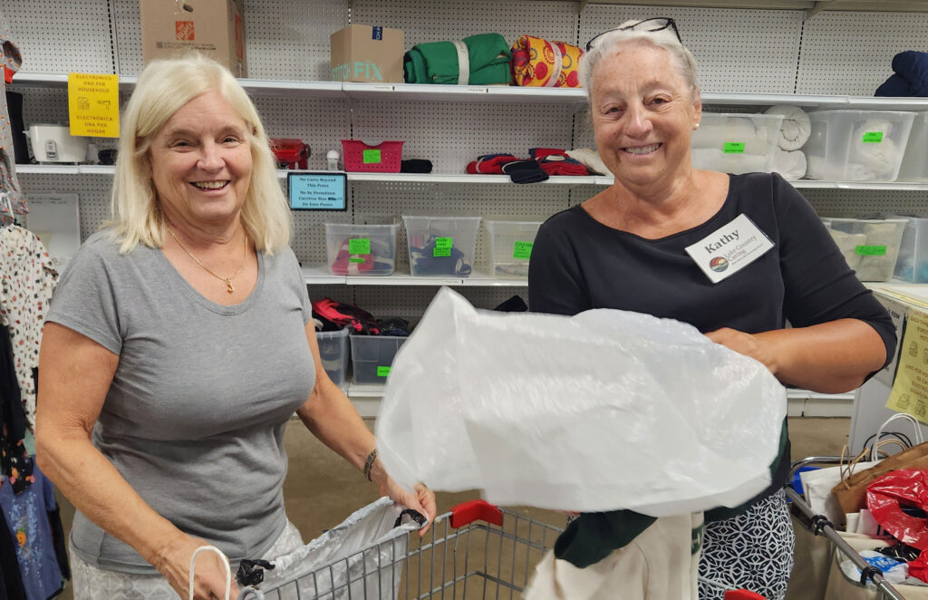 Two smiling older women sort clothing donations in a thrift store. One holds a large white plastic bag, while the other stands by a shopping cart. Shelves with bins and items are visible in the background.