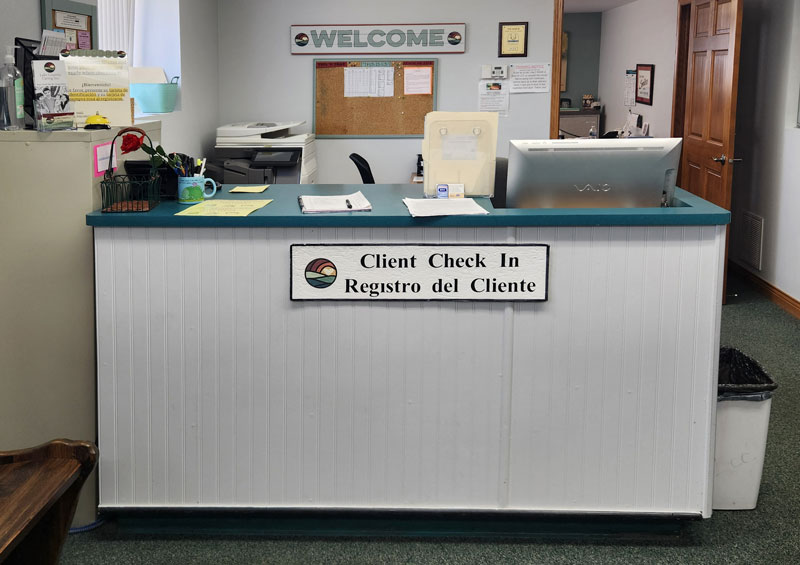A reception desk with a sign reading Client Check In / Registro del Cliente in English and Spanish. Behind the desk are office supplies, a computer, and a bulletin board. A WELCOME sign hangs on the wall.
